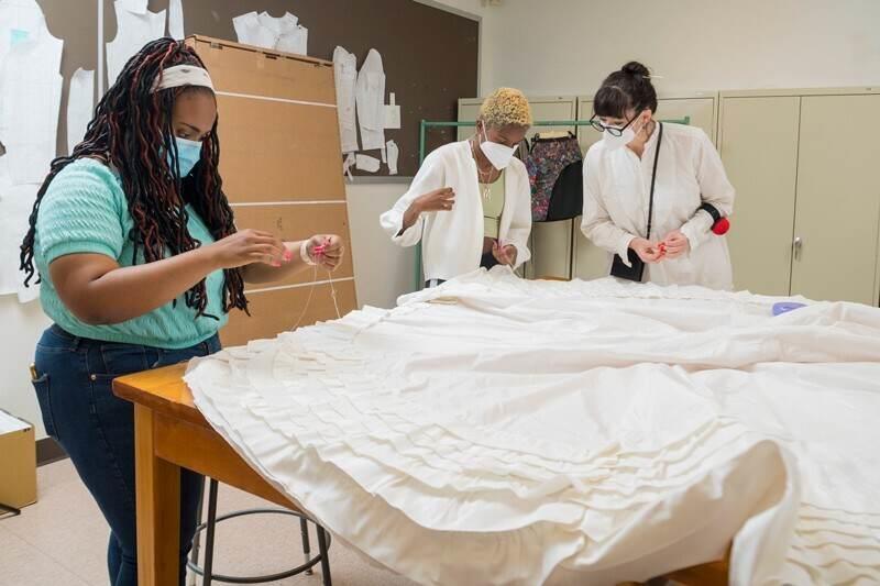 three people hand sewing white fabrice laid out on a table